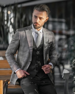 A man dressed in a stylish checkered blazer and vest stands confidently with one hand in his pocket. He is in a modern, possibly indoor setting with blurred background elements such as chairs and tables. The lighting casts shadows and highlights on his face and suit.