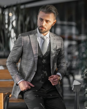 A man dressed in a stylish checkered blazer and vest stands confidently with one hand in his pocket. He is in a modern, possibly indoor setting with blurred background elements such as chairs and tables. The lighting casts shadows and highlights on his face and suit.