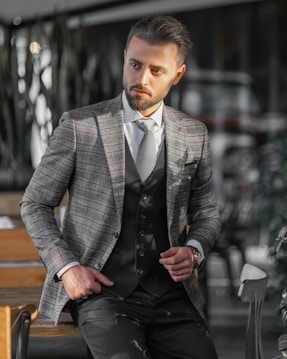A man dressed in a stylish checkered blazer and vest stands confidently with one hand in his pocket. He is in a modern, possibly indoor setting with blurred background elements such as chairs and tables. The lighting casts shadows and highlights on his face and suit.
