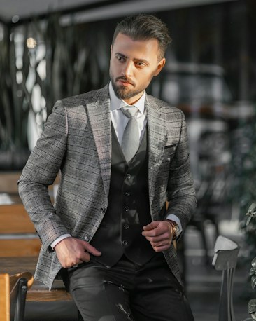 A man dressed in a stylish checkered blazer and vest stands confidently with one hand in his pocket. He is in a modern, possibly indoor setting with blurred background elements such as chairs and tables. The lighting casts shadows and highlights on his face and suit.