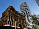 A historic building with ornate architecture stands prominently, characterized by its intricate arches and detailed facade. Adjacent to it is a modern skyscraper with a striped pattern of alternating dark and light glass, stretching towards the clear blue sky. Lush green trees are visible, suggesting an urban setting with a mix of old and new architectural styles.
