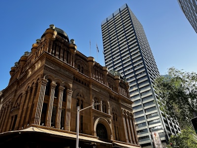 A historic building with ornate architecture stands prominently, characterized by its intricate arches and detailed facade. Adjacent to it is a modern skyscraper with a striped pattern of alternating dark and light glass, stretching towards the clear blue sky. Lush green trees are visible, suggesting an urban setting with a mix of old and new architectural styles.