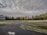 Overcast sky with thick clouds stretches across a rural landscape. A paved road intersects a grassy field lined with dense trees. A sign for New Life Christian Center Ministries is on the left, beside the road, indicating service times.