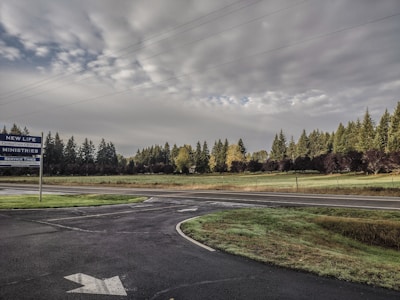 Overcast sky with thick clouds stretches across a rural landscape. A paved road intersects a grassy field lined with dense trees. A sign for New Life Christian Center Ministries is on the left, beside the road, indicating service times.