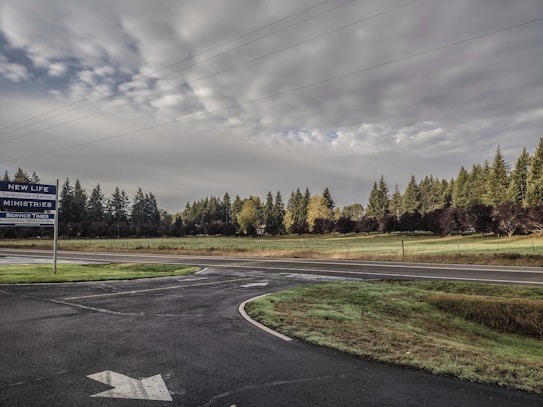 Overcast sky with thick clouds stretches across a rural landscape. A paved road intersects a grassy field lined with dense trees. A sign for New Life Christian Center Ministries is on the left, beside the road, indicating service times.