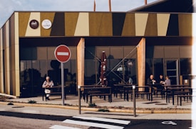 A modern cafe with a geometric facade featuring olive and beige tones. Outdoor seating includes wooden tables and chairs. A 'no entry' sign is visible in the foreground and several people are seated at the tables, engaged in conversation or using their phones. The scene has a quiet, relaxed atmosphere.