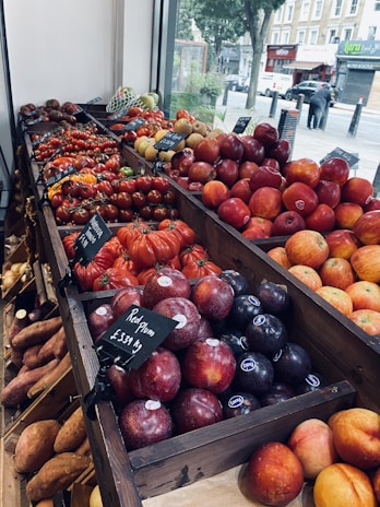 A market display showcasing a variety of fresh fruits and vegetables arranged neatly in wooden crates. Red plums and tomatoes are prominently featured, with price tags visible. The display is next to a large window, revealing an outdoor street scene with trees and shops in the background.