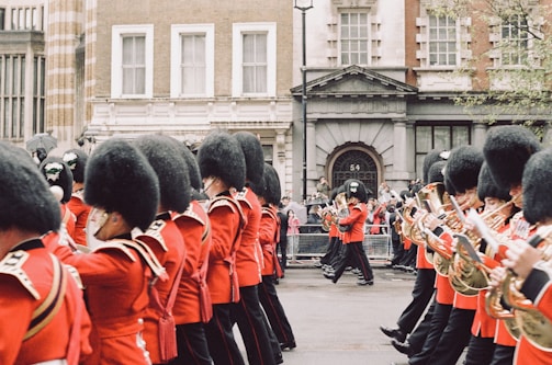 A parade of uniformed musicians marching along a street. They wear traditional red uniforms with black hats, and each holds a musical instrument, primarily brass. The backdrop includes historic buildings and a small gathering of spectators.