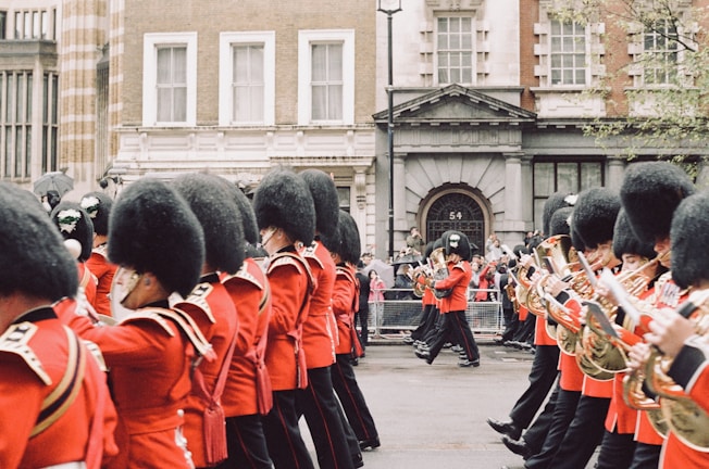 A parade of uniformed musicians marching along a street. They wear traditional red uniforms with black hats, and each holds a musical instrument, primarily brass. The backdrop includes historic buildings and a small gathering of spectators.