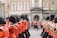 A parade of uniformed musicians marching along a street. They wear traditional red uniforms with black hats, and each holds a musical instrument, primarily brass. The backdrop includes historic buildings and a small gathering of spectators.