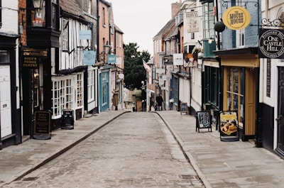 A narrow, cobblestone street lined with quaint shops and businesses. The buildings have a variety of colorful signs and traditional architectural features. There are outdoor signage boards on the sidewalk, and potted plants hanging from some shopfronts. The street is empty, adding to the historical and charming atmosphere of the scene.