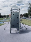 A playground scene featuring a climbing structure with a vertical wall fitted with holds, surrounded by ropes and metal bars. In the background, a slide and other play equipment are visible. The area is enclosed with a metal fence and bordered by grass and trees under a partly cloudy sky.