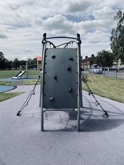 A playground scene featuring a climbing structure with a vertical wall fitted with holds, surrounded by ropes and metal bars. In the background, a slide and other play equipment are visible. The area is enclosed with a metal fence and bordered by grass and trees under a partly cloudy sky.