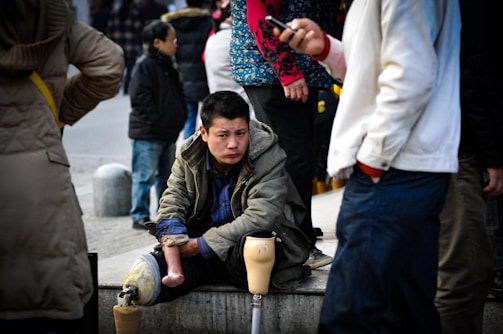 A man with prosthetic legs sits on a ledge in an urban setting, surrounded by people passing by. He wears a heavy jacket and appears contemplative. Various individuals in casual attire walk around him, some focused on their phones.