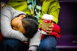 A young child is asleep with their head resting on their arm and lap. They are wearing a gray hoodie. An adult, dressed in a bright green sweater and multicolored scarf, holds a paper coffee cup with a red sleeve labeled 'SHARE'. The setting appears to be a public place, possibly a subway or bus.