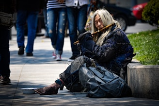 A person with long hair wearing tattered clothing is sitting on the street near a patch of grass. The person is holding a flute and appears to be playing it. There is a bowl on the ground in front of the person, likely used for collecting donations. In the background, pedestrians are walking by, and parked cars are visible.