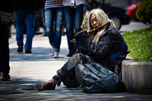 A person with long hair wearing tattered clothing is sitting on the street near a patch of grass. The person is holding a flute and appears to be playing it. There is a bowl on the ground in front of the person, likely used for collecting donations. In the background, pedestrians are walking by, and parked cars are visible.