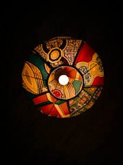 A colorful, circular stained-glass lampshade is illuminated by a central light. The lampshade features intricate geometric patterns and bold colors, including red, green, yellow, and blue, forming a vibrant and visually striking design. The dark background enhances the vividness of the colors and patterns.