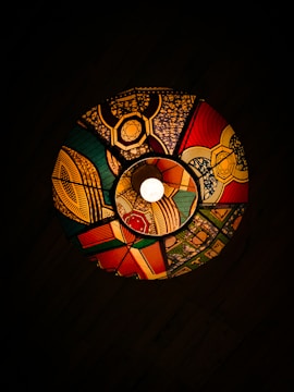 A colorful, circular stained-glass lampshade is illuminated by a central light. The lampshade features intricate geometric patterns and bold colors, including red, green, yellow, and blue, forming a vibrant and visually striking design. The dark background enhances the vividness of the colors and patterns.