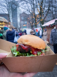 A close-up of a hamburger with lettuce and ketchup, served in a brown cardboard box against a blurred background of an outdoor market setting. People are casually walking around, and trees with decorative lights add to the festive atmosphere.