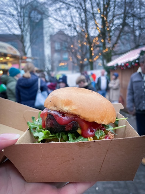 A close-up of a hamburger with lettuce and ketchup, served in a brown cardboard box against a blurred background of an outdoor market setting. People are casually walking around, and trees with decorative lights add to the festive atmosphere.