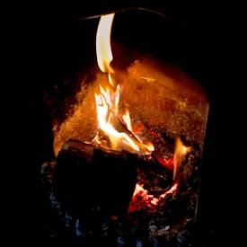 A wood log burning with bright orange and yellow flames, casting a warm glow in a dimly lit space.