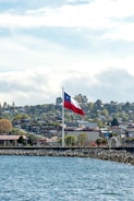 A large flag waves prominently above a coastal area with a rocky shoreline. The scene in the background features a hilly townscape with residential buildings and trees under a partly cloudy sky.