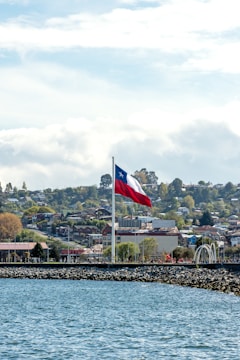 A large flag waves prominently above a coastal area with a rocky shoreline. The scene in the background features a hilly townscape with residential buildings and trees under a partly cloudy sky.