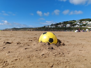 A yellow and black soccer ball partially buried in the sand on a beach with a clear blue sky overhead. In the background, there are scattered people, and houses are visible on a hillside.