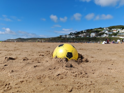 A yellow and black soccer ball partially buried in the sand on a beach with a clear blue sky overhead. In the background, there are scattered people, and houses are visible on a hillside.