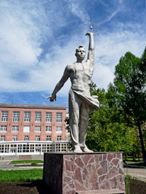 A tall statue of a man stands outdoors on a stone pedestal. The figure has one arm raised holding a model of an atom, possibly symbolizing scientific achievement. Behind the statue, there is a red-brick building with several windows and a visible entrance. The scene includes lush green trees and a partly cloudy sky, suggesting a pleasant day.