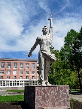 A tall statue of a man stands outdoors on a stone pedestal. The figure has one arm raised holding a model of an atom, possibly symbolizing scientific achievement. Behind the statue, there is a red-brick building with several windows and a visible entrance. The scene includes lush green trees and a partly cloudy sky, suggesting a pleasant day.