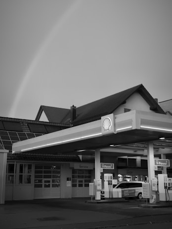 A black and white image of a gas station featuring a covered fueling area with multiple fuel pumps. The station has a prominent canopy displaying the Shell logo. In the background, there are buildings with pitched roofs and a visible garage or service area. A car is parked near the pumps, and a rainbow is faintly visible in the sky.