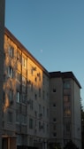 A multi-story residential building with a brick facade is illuminated by the warm light of the setting sun, casting shadows on the wall. The sky is clear with a visible crescent moon.