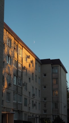 A multi-story residential building with a brick facade is illuminated by the warm light of the setting sun, casting shadows on the wall. The sky is clear with a visible crescent moon.