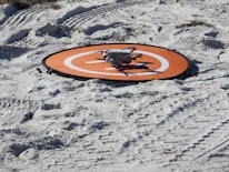 A small drone rests on an orange landing pad placed on sandy terrain with visible tire tracks. The sand appears white with a few scattered debris, and the layout suggests preparation for drone operation.