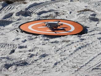 A small drone rests on an orange landing pad placed on sandy terrain with visible tire tracks. The sand appears white with a few scattered debris, and the layout suggests preparation for drone operation.