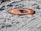A small drone rests on an orange landing pad placed on sandy terrain with visible tire tracks. The sand appears white with a few scattered debris, and the layout suggests preparation for drone operation.