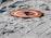A small drone rests on an orange landing pad placed on sandy terrain with visible tire tracks. The sand appears white with a few scattered debris, and the layout suggests preparation for drone operation.