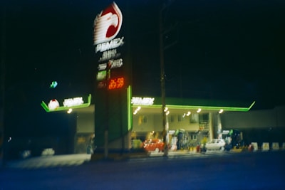 A night-time scene of a gas station with bright green and white lighting. The gas station has a prominently displayed sign with red, white, and green elements, and a visible logo at the top. The station features a lit canopy over the pumps and several cars parked in front. The overall scene is slightly blurry and dark, contributing to an unclear but vibrant look.