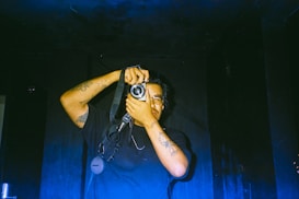 A person is holding a camera up to their face, capturing a photo. The individual has tattoos on their arms and is wearing glasses. The background is dark, creating a high contrast with the subject.