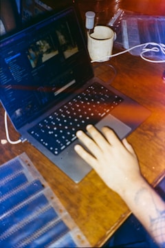 A hand is placed on the keyboard of a laptop displaying photo editing software. There is a mug and some film strips on a wooden table, along with a charger connected to the laptop.