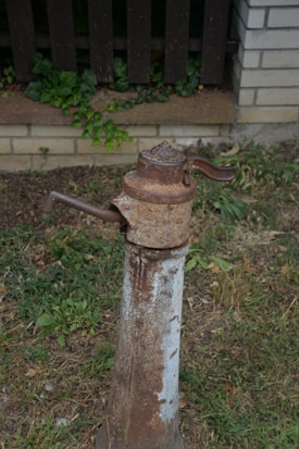 An old, rusty water pump is situated in a grassy area, with a wooden fence and bricks in the background. The pump has a weathered appearance with visible corrosion and a handle on the side.