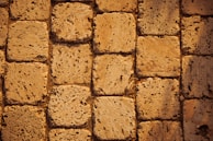 A close-up view of a textured stone pavement. The stones are rectangular and laid out in a neat pattern. The surface appears rough and porous, suggesting wear and natural erosion over time. The color is a warm beige, adding a rustic vibe.