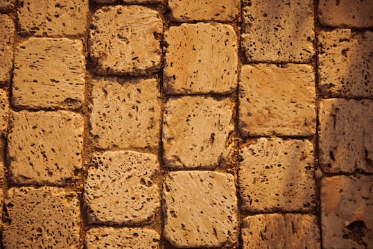 A close-up view of a textured stone pavement. The stones are rectangular and laid out in a neat pattern. The surface appears rough and porous, suggesting wear and natural erosion over time. The color is a warm beige, adding a rustic vibe.