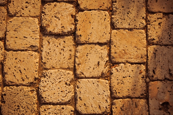 A close-up view of a textured stone pavement. The stones are rectangular and laid out in a neat pattern. The surface appears rough and porous, suggesting wear and natural erosion over time. The color is a warm beige, adding a rustic vibe.