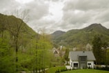 A modern house with a grey roof is situated in a lush green valley surrounded by dense trees and hills. In the background, taller mountains rise under a cloudy sky. A small path leads to the house, and a few more buildings with steep roofs can be seen in the distance.