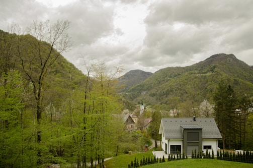 A modern house with a grey roof is situated in a lush green valley surrounded by dense trees and hills. In the background, taller mountains rise under a cloudy sky. A small path leads to the house, and a few more buildings with steep roofs can be seen in the distance.