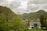 A modern house with a grey roof is situated in a lush green valley surrounded by dense trees and hills. In the background, taller mountains rise under a cloudy sky. A small path leads to the house, and a few more buildings with steep roofs can be seen in the distance.