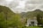 A modern house with a grey roof is situated in a lush green valley surrounded by dense trees and hills. In the background, taller mountains rise under a cloudy sky. A small path leads to the house, and a few more buildings with steep roofs can be seen in the distance.
